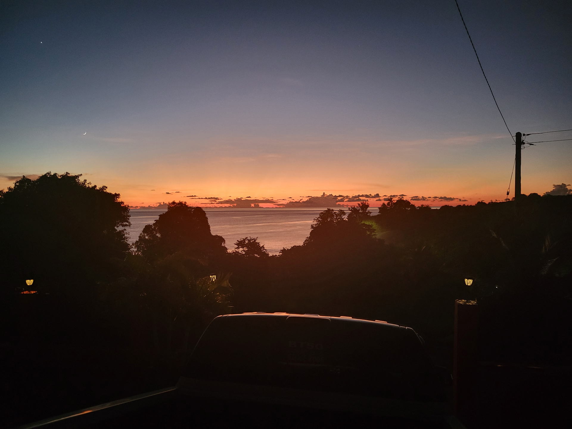 Twilight sea horizon with orange glow, crescent moon, and silhouettes of trees